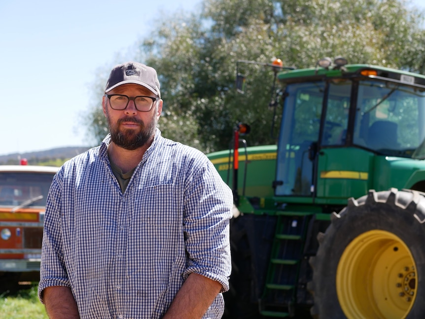A man in checkered shirt on a farm standing in front of a green tractor