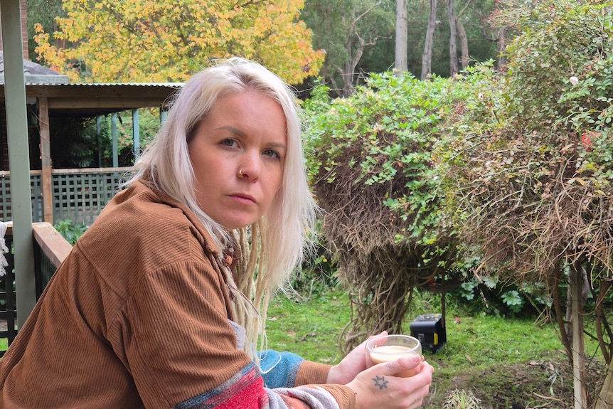 A young woman with pale blonde hair in a brown shirt holds a coffee while leaning over a balcony on a deck.