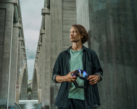 A man with reddish chin-length hair in casual clothing holding large ski goggles on a concrete bridge