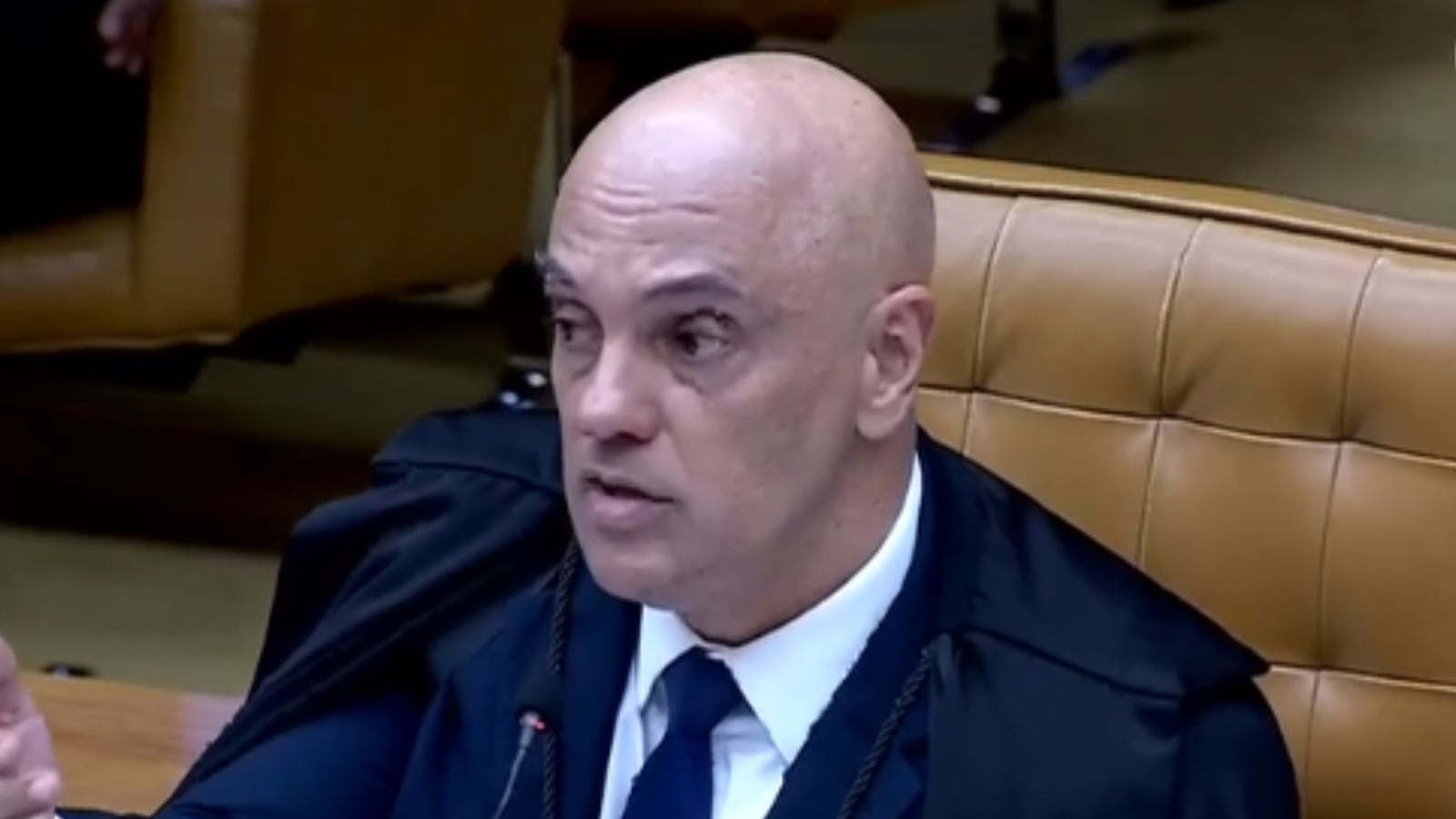Um homem calvo, vestindo uma toga preta e uma camisa branca com gravata, está falando em um ambiente formal
