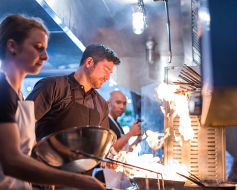 Chefs work at a flaming grill in a restaurant kitchen, one holding a metal bowl